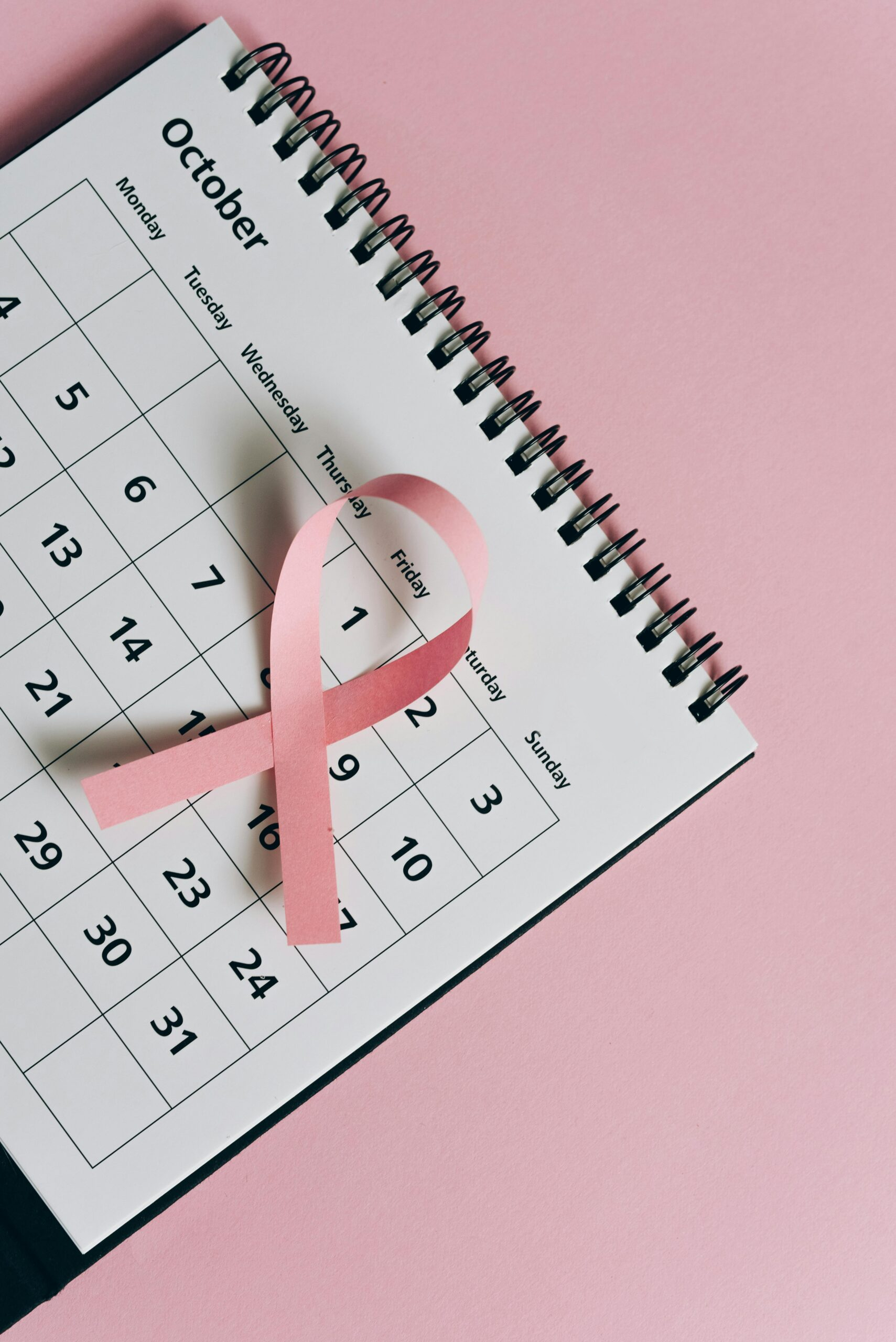 An October calendar lies at an angle on a pink background. A breast cancer awareness ribbon is on the calendar